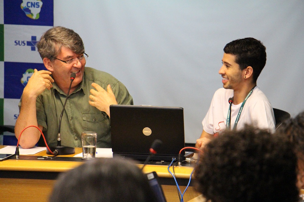 #PraCegoVer: Na imagem, Ronald dos Santos e Douglas Pereira aparecem sorrindo, sentados à mesa do plenário do CNS.
